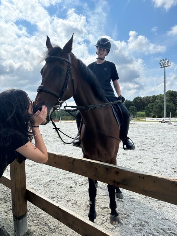 A rider on a bay horse at a wooden fence while a friend strokes its nose